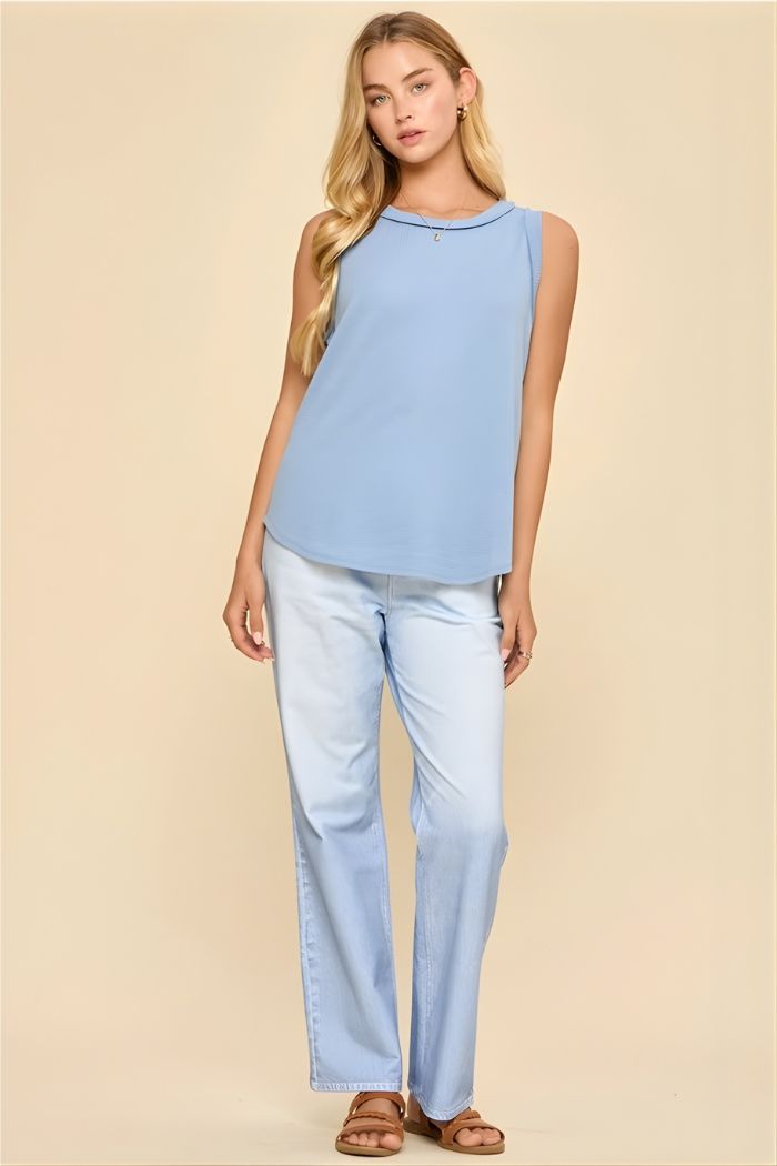 A woman with long, wavy blonde hair stands against a beige background wearing a light blue Basic Knit Tank Top, matching jeans, and brown sandals. She has a relaxed, neutral expression.
