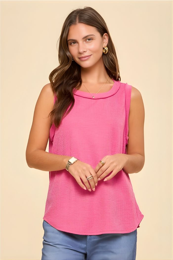A woman with long brown hair wears the Basic Knit Tank Top in textured pink, gold hoop earrings, layered necklaces, rings, and a watch while smiling gently against a plain beige background.
