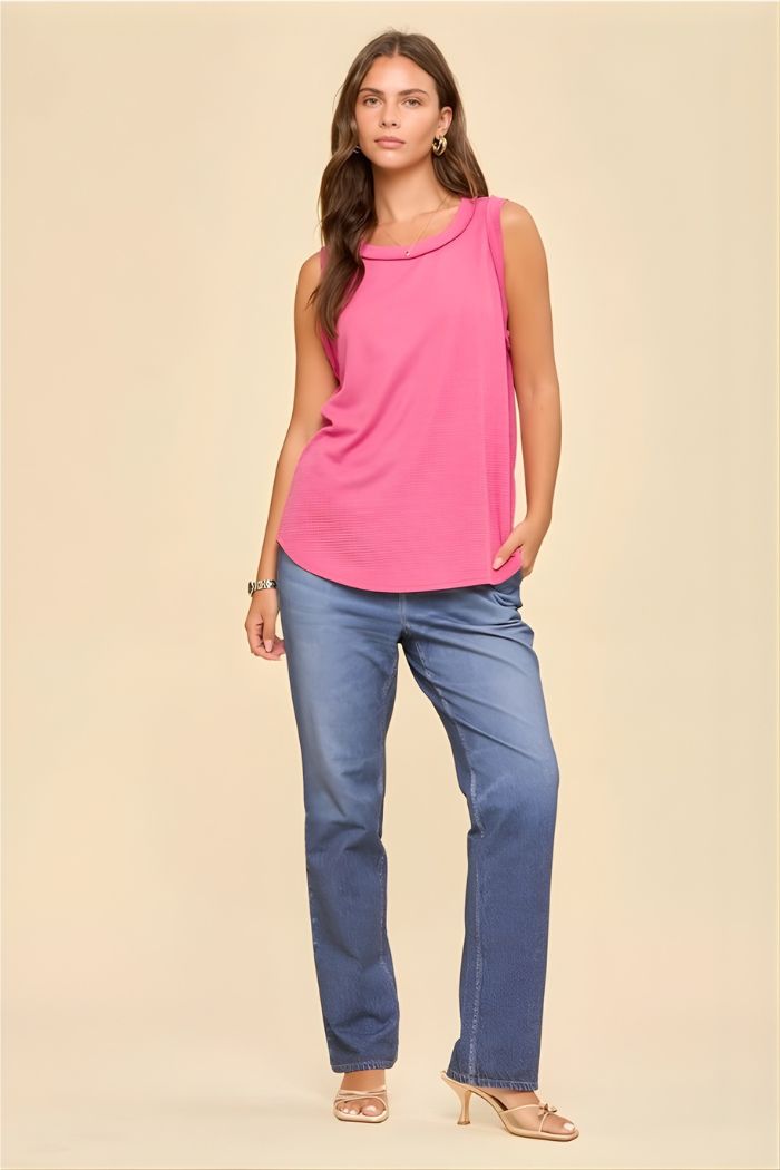 A woman stands against a beige background wearing a Basic Knit Tank Top in pink, blue straight-leg jeans, beige heeled sandals, hoop earrings, and a bracelet. She has long brown hair.