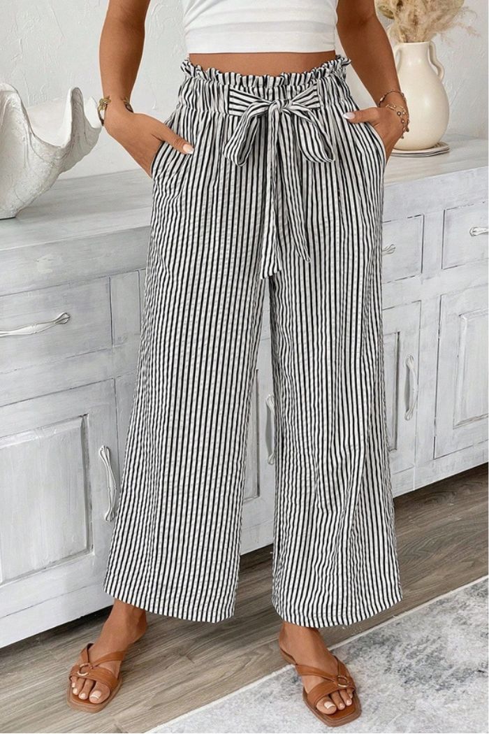 A person stands indoors with hands in pockets, wearing Striped Paperbag Waist Pants, a white crop top, and brown sandals.