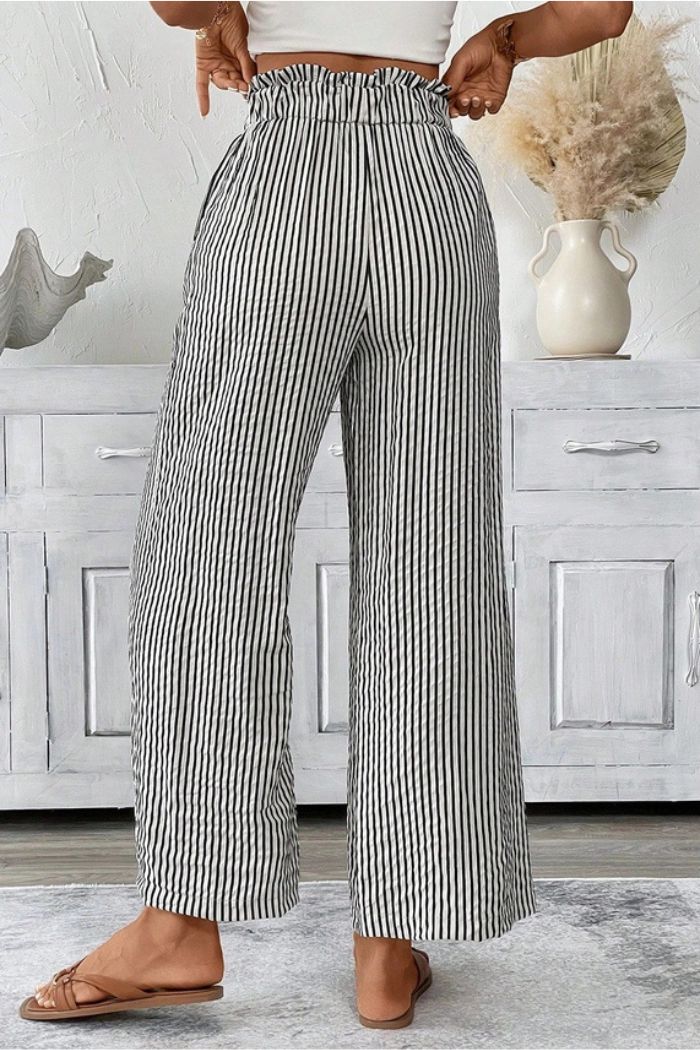 A woman stands indoors wearing Striped Paperbag Waist Pants and brown flat sandals, facing away with her hands on her waist. A white vase with dried flowers is on a cabinet in the background.