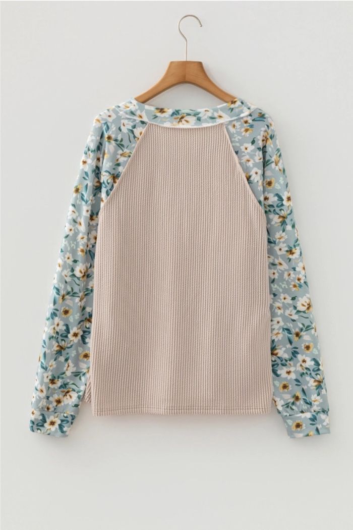 The Floral Contrast Ribbed Top hangs on a wooden hanger, showcasing a textured beige ribbed knit back and blue sleeves with a white and yellow floral print, set against a plain white background.