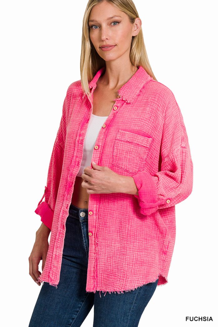 A woman wears a bright fuchsia Washed Double Gauze Shirt by Zenana over a white crop top and jeans, standing with one hand touching the shirt. The oversized shirt features a chest pocket and rolled-up sleeves.