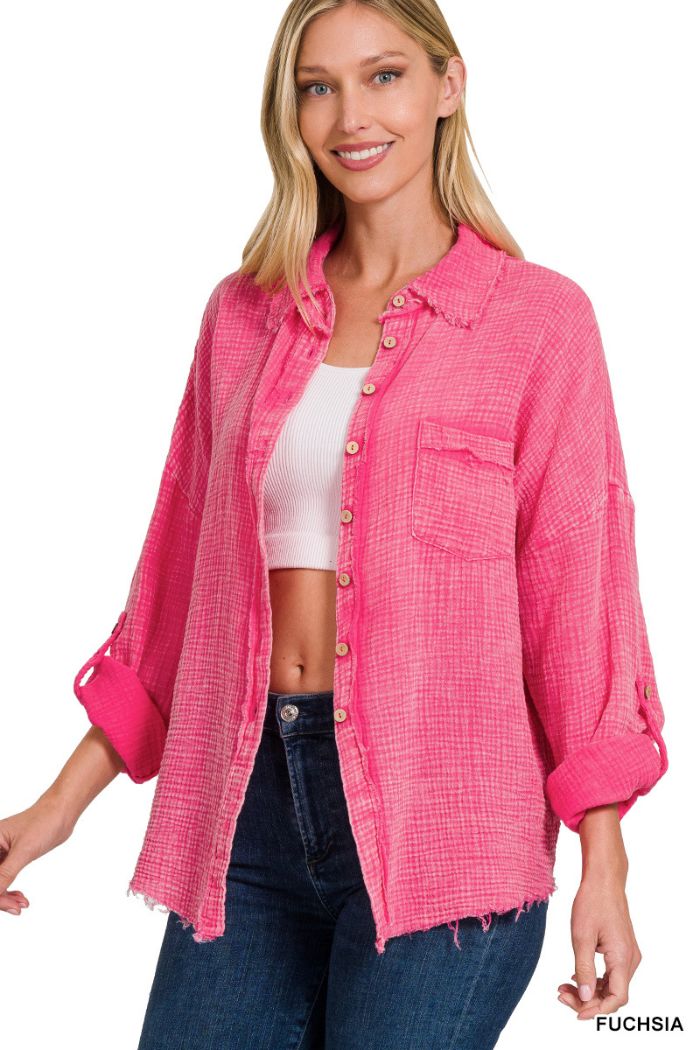 A woman with long blonde hair smiles as she wears a Washed Double Gauze Shirt by Zenana in fuchsia over a white crop top and dark jeans. The textured, oversized shirt features frayed edges, rolled sleeves, and a chest pocket.