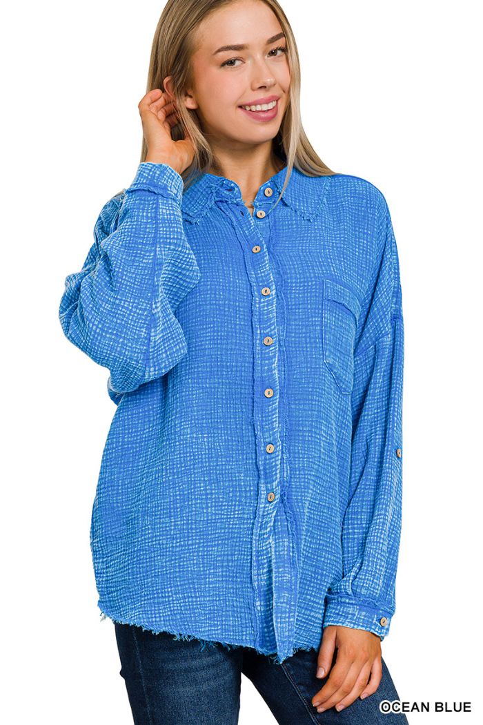 A smiling woman wears a Zenana Washed Double Gauze Shirt in textured blue with long sleeves and dark jeans, posing with one hand touching her hair.