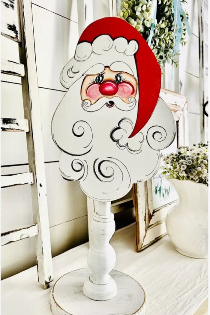 The Christmas Wood Topper features a cheerful, vintage-style Santa face with a red hat and white beard, hand-painted on wood and set on a white stand—adding rustic charm to your festive holiday décor.