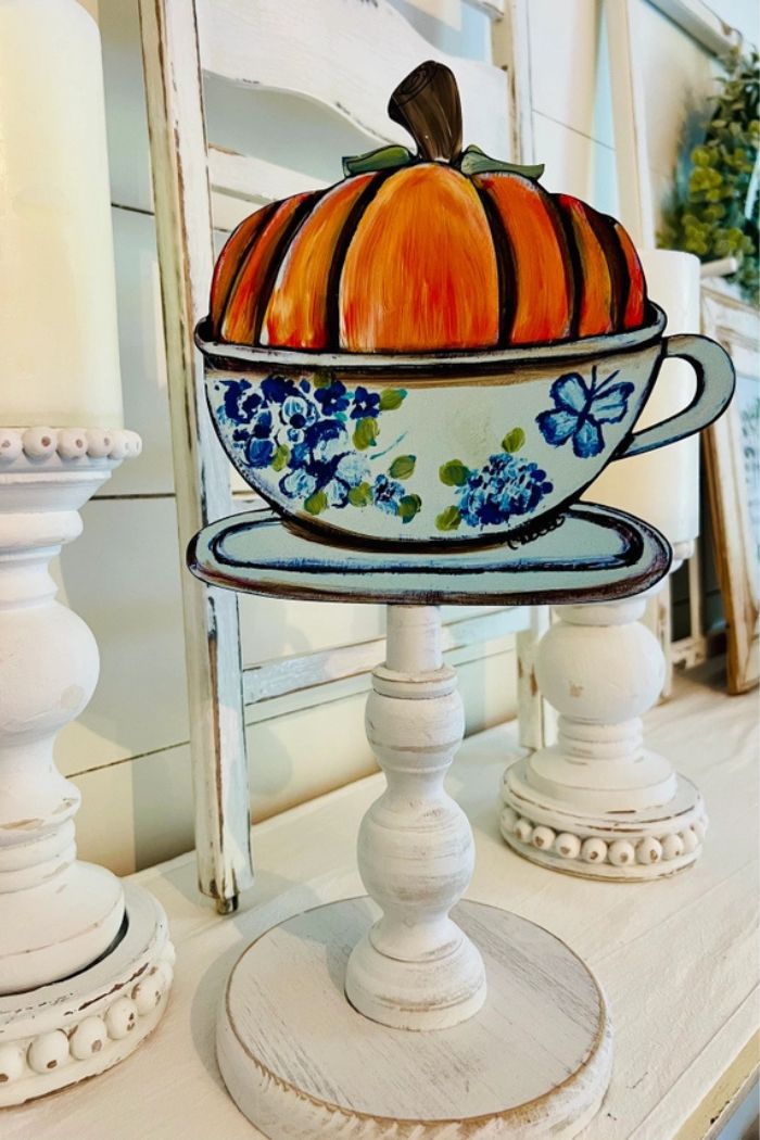Fall Wood Toppers feature a painted teacup and saucer with blue floral designs, topped by a hand-painted wooden pumpkin on a white stand—perfect for farmhouse seasonal displays and autumn home decor.