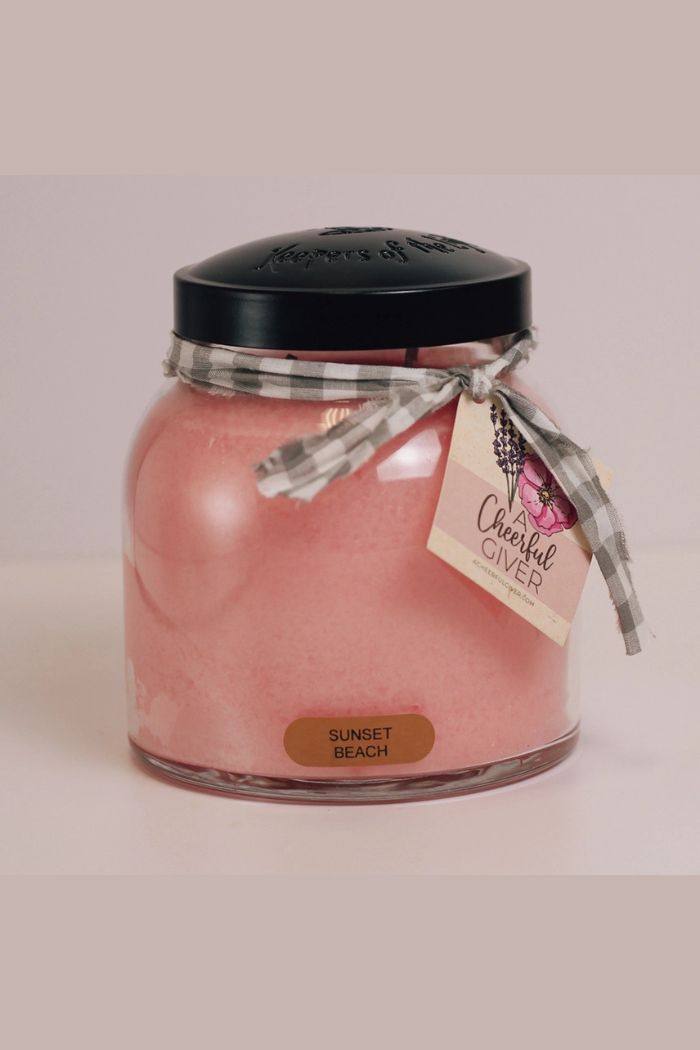 The Papa 34oz Candle is a large scented candle in a glass jar with a black lid, adorned with a gray plaid ribbon and gift tag. Its label reads "Sunset Beach," making it the perfect decorative addition to any room.