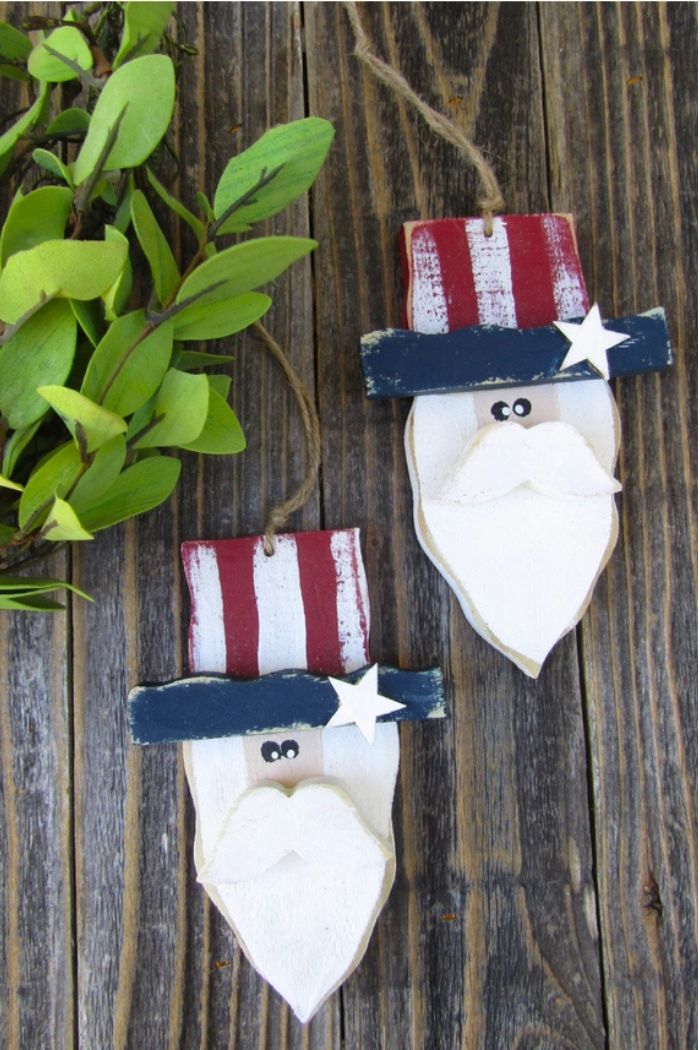 Two Uncle Sam Wood Ornaments, featuring white beards, blue star-spangled hats, and red stripes, hang by twine on a rustic wood surface beside green leaves—perfect patriotic décor for your home.