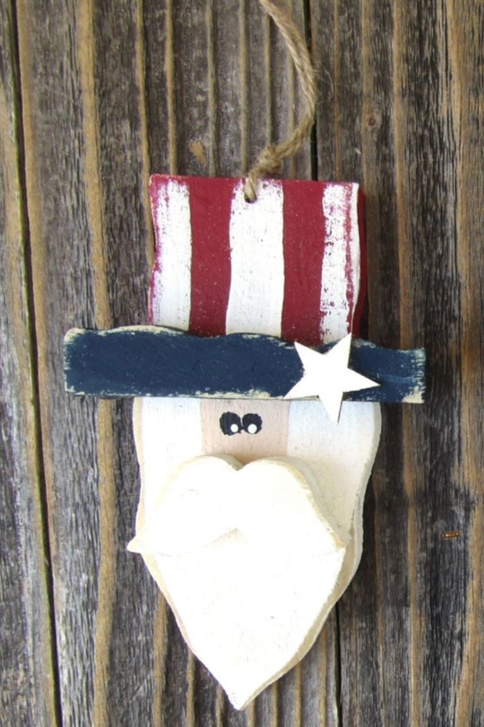 The Uncle Sam Wood Ornament, featuring a white-bearded face with a red and white striped hat, blue band, and white star, hangs on rustic wood—a perfect piece for Americana décor.