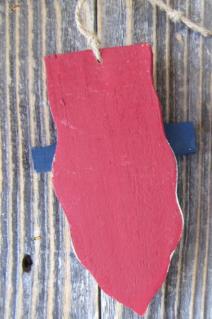 The Uncle Sam Wood Ornament, a red shield-shaped patriotic design, hangs by a twine loop on weathered wood, with two small dark blue tabs peeking behind the sides.