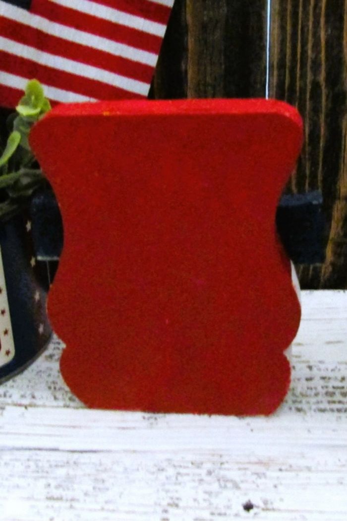 The Rustic Uncle Sam Decor, a small red wooden block with a curved shape, rests on a white surface near a potted plant and an American flag, adding charming farmhouse patriotic style to your space.
