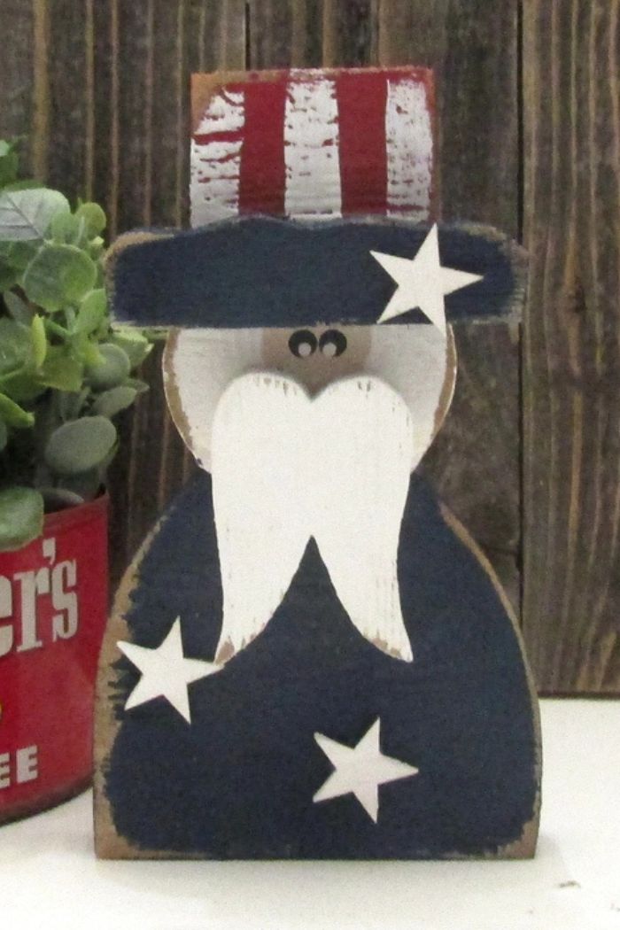 The Patriotic Uncle Sam Decor features a wooden figure with a white beard, striped hat, and starry blue coat, bringing classic Americana style to your space when displayed on a rustic background beside a potted plant.