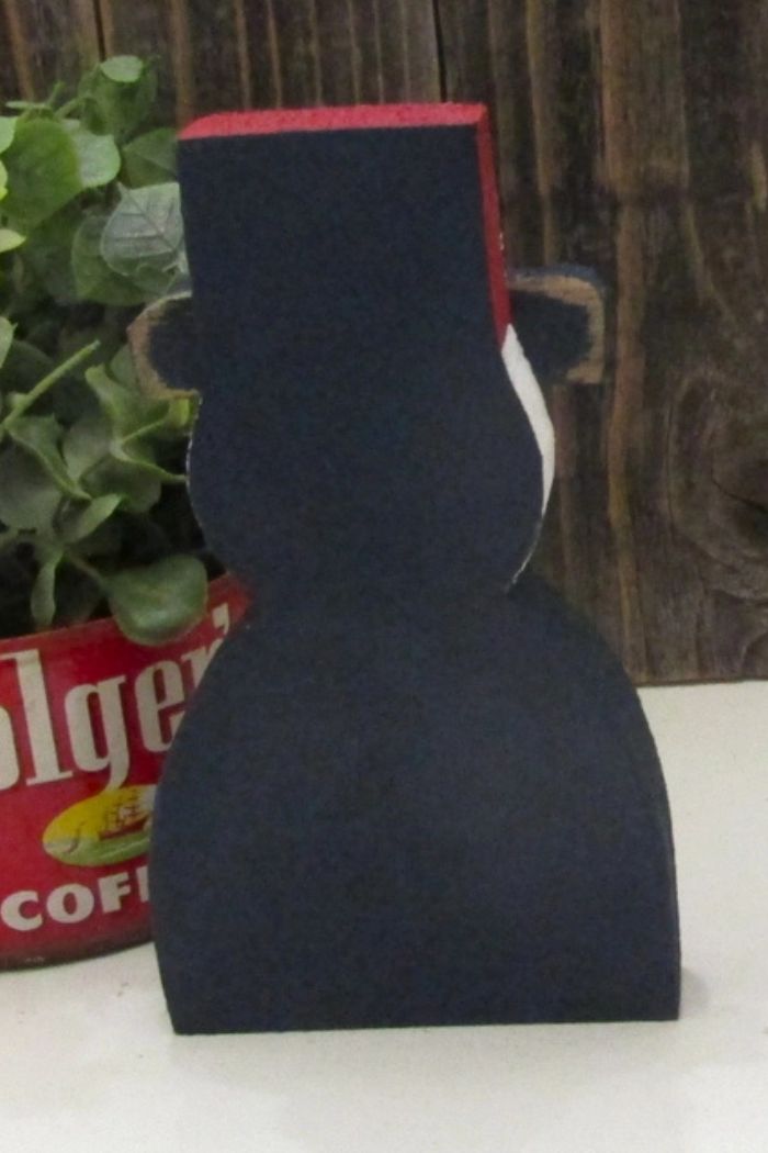 The Patriotic Uncle Sam Decor, painted black with a red top hat, stands before rustic wood and a Folgers tin of greenery. The snowman's unseen features add to this unique Americana-inspired display.