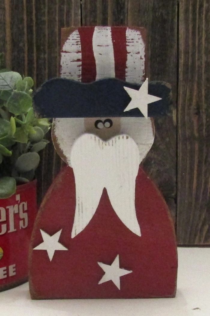 The Patriotic Uncle Sam Decor features a wooden figure in vibrant red, white, and blue with white stars. A green plant in a red container sits beside it, all displayed against rustic wooden plank backgrounds.