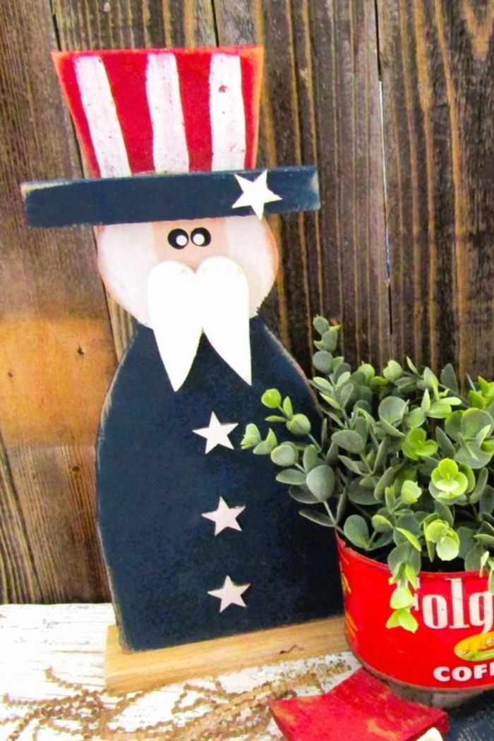 The Large Uncle Sam Decor features a rustic wood figure with a white beard, blue coat, and striped hat. It stands beside a red Folgers coffee can planter with greenery—perfect for patriotic farmhouse decor against a wooden backdrop.