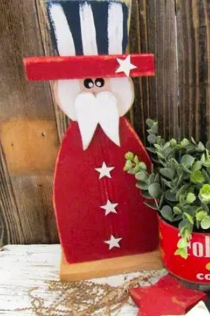 The Large Uncle Sam Decor, featuring a rustic wood figure in a red coat and tall hat beside a red Folgers coffee can with a green potted plant, creates charming patriotic farmhouse decor on a weathered wooden surface.