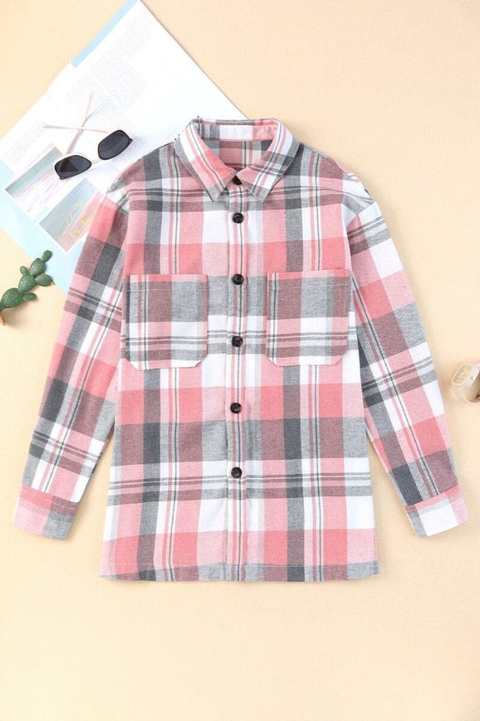 The Plaid Button Up Shirt in pink, gray, and white is laid flat on a beige surface beside sunglasses, a magazine, a small cactus, and a cork. It features two chest pockets and classic flannel style.