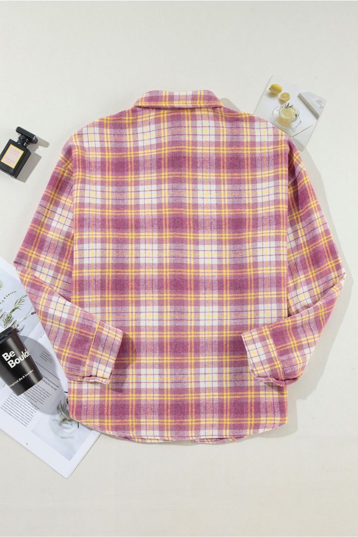 The Pink Plaid Shacket is laid flat on a light surface with magazines, a perfume bottle, and a glass of lemon water nearby. The back of the shacket is shown.