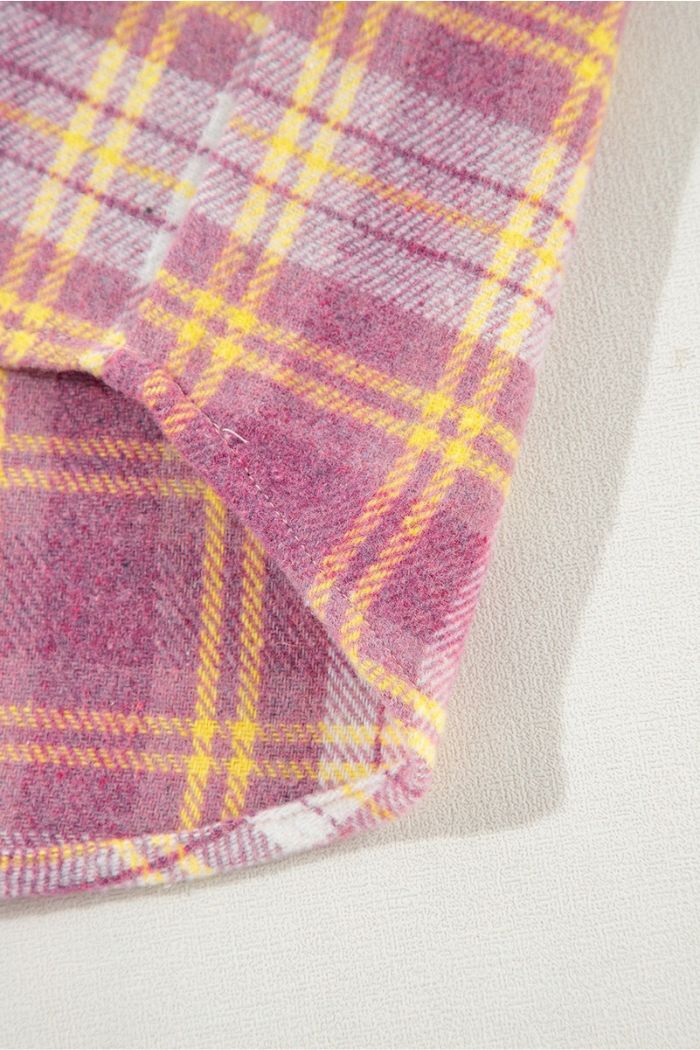 A close-up of the Pink Plaid Shacket shows its soft pink and yellow plaid fabric with visible stitching, folded at the corner, highlighting its cozy texture as a stylish women's transitional jacket on a light textured surface.