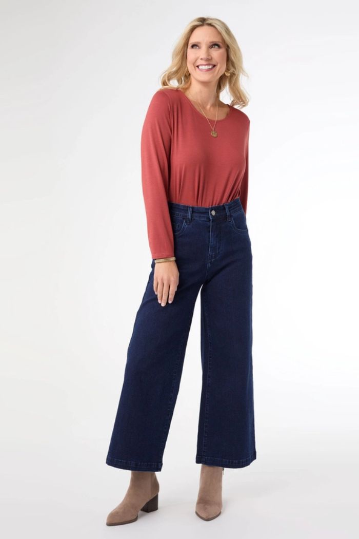 A woman with blonde hair smiles in a rust long-sleeve top, dark blue Everstretch Cropped Wide Jeans, tan heeled ankle boots, and a necklace, standing against a plain white background.