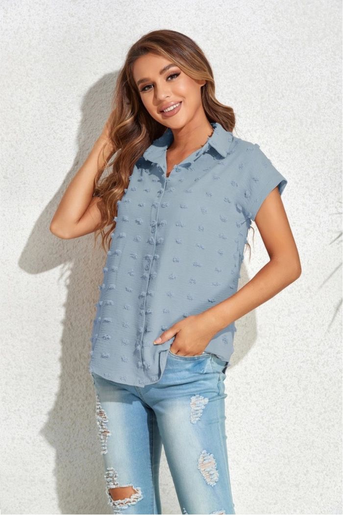 A woman with long, wavy hair smiles while standing against a light textured wall, wearing a Textured Button Blouse paired with light blue ripped jeans and posing with one hand in her pocket.