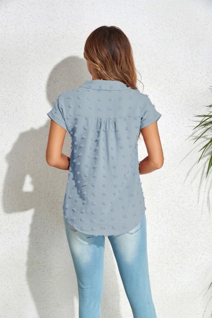 A woman with long brown hair stands facing a white textured wall, wearing a light blue Textured Button Blouse with cap sleeves and faded blue jeans. She is shown from the back, with green plant leaves visible on the right side.