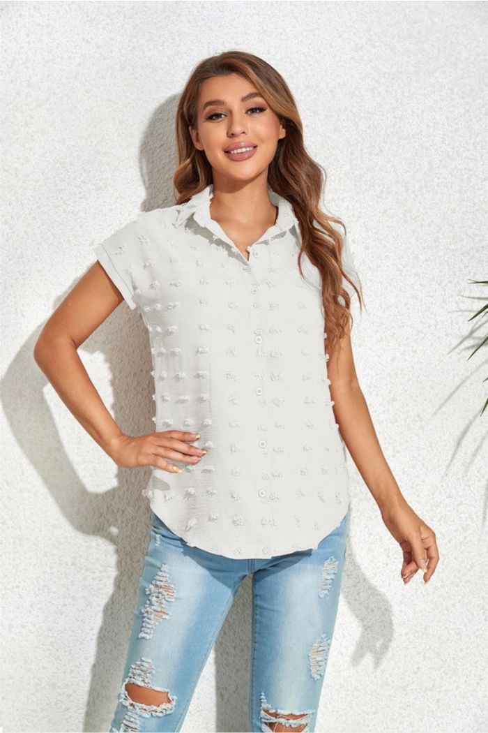 A woman smiles against a light textured wall, wearing a white Textured Button Blouse with short sleeves and light blue ripped jeans. One hand is on her hip, the other by her side.