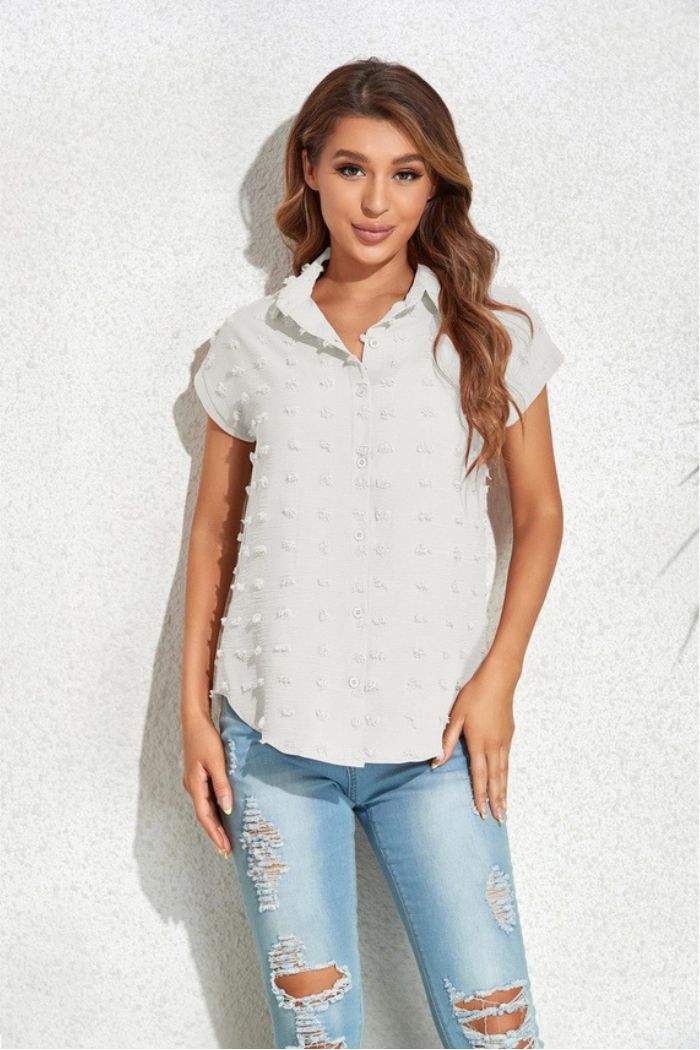 A woman with long, wavy hair smiles gently at the camera, standing against a light textured wall in a Textured Button Blouse with short sleeves and ripped light blue jeans.