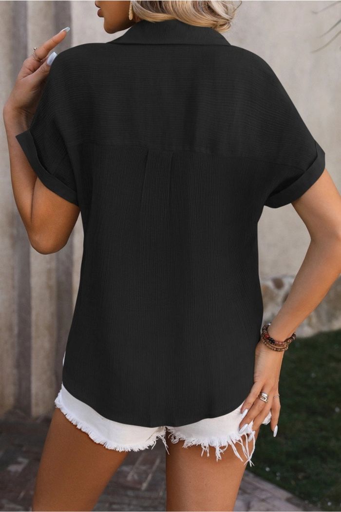 A person stands outdoors with their back to the camera, wearing an Open Front Black Blouse with short sleeves and frayed white denim shorts, a bracelet on one wrist, and touching their hair with one hand.