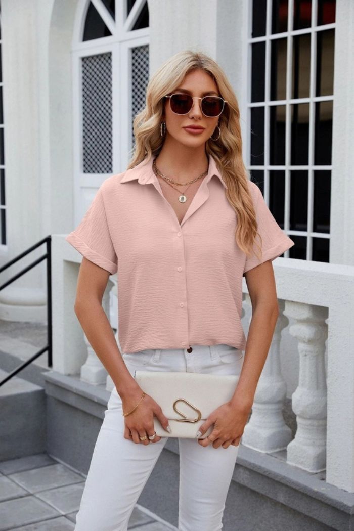 A woman with long blonde hair wearing sunglasses, white pants, and a light pink Button Down Blouse stands outdoors holding a white clutch in front of a white building with arched windows and a railing.