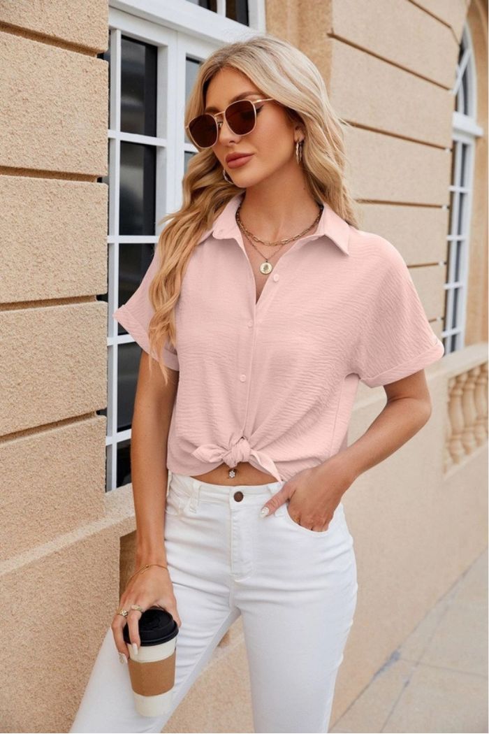 A woman with long blonde hair wearing sunglasses, a light pink Button Down Blouse tied at the waist and white pants stands outside holding a coffee cup in front of a beige building with arched windows.