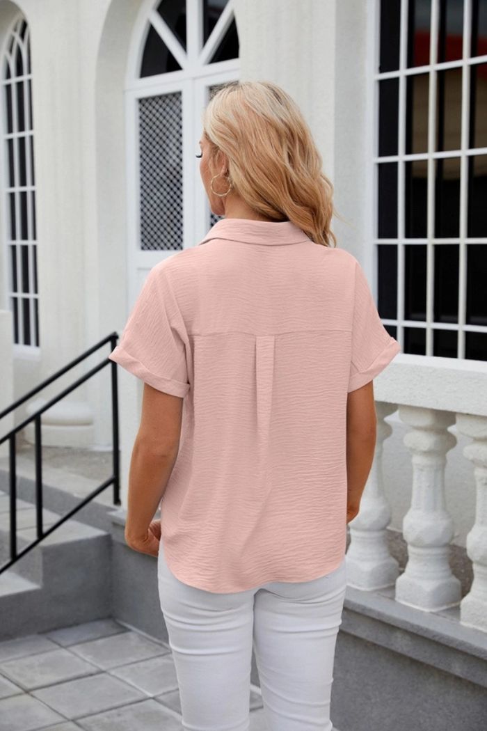 A woman with blonde hair, seen from behind, wears a light pink Button Down Blouse and white pants while standing on outdoor steps near a white building with arched windows and railings.