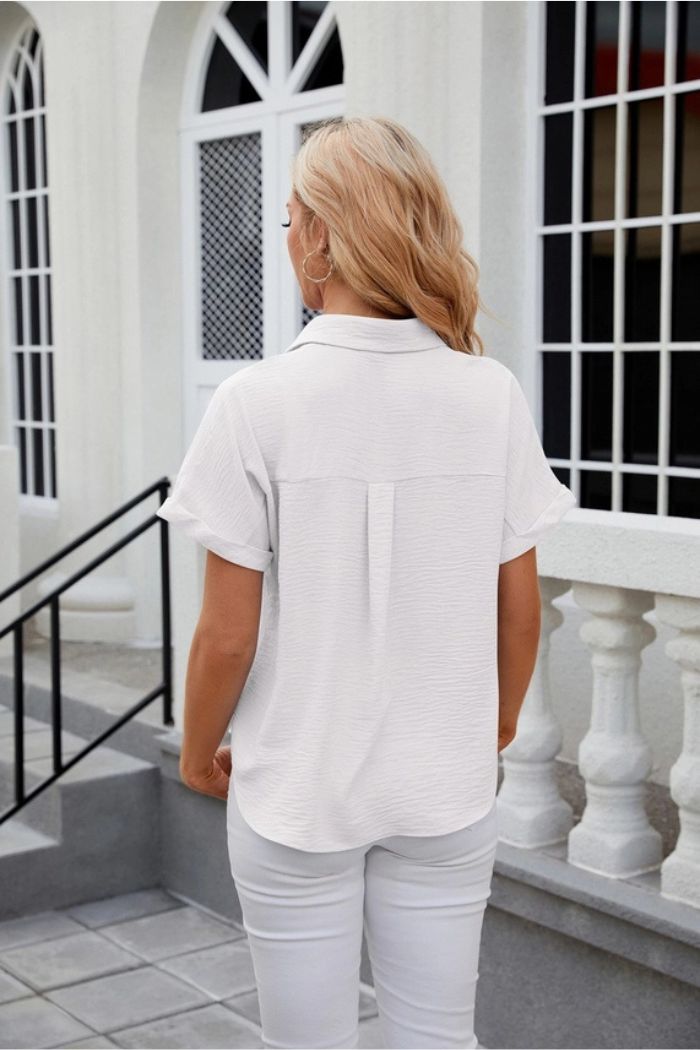A woman with blonde hair, dressed in a white Button Down Blouse and white pants, stands with her back to the camera near white balustrades and windows.