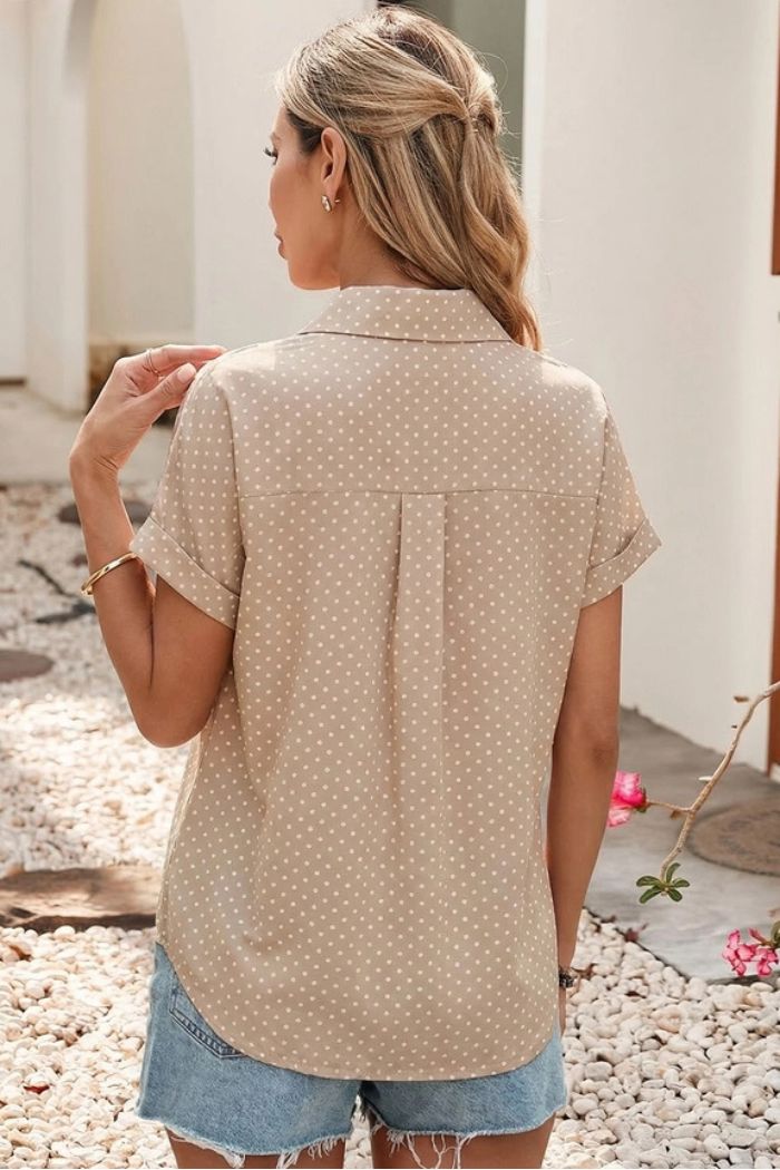 A woman with long, light brown hair stands outdoors, facing away, wearing the Dotted Cuff Sleeve Shirt in beige and light blue denim shorts, amidst white walls and a gravel path dotted with pink flowers.