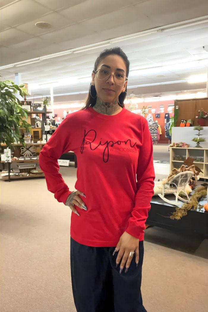 A woman with glasses and tattoos smiles indoors, posing with one hand on her hip in a store filled with plants. She wears the Ripon Wisconsin Graphic Tee, featuring a classic crew-neck design and bold graphics.