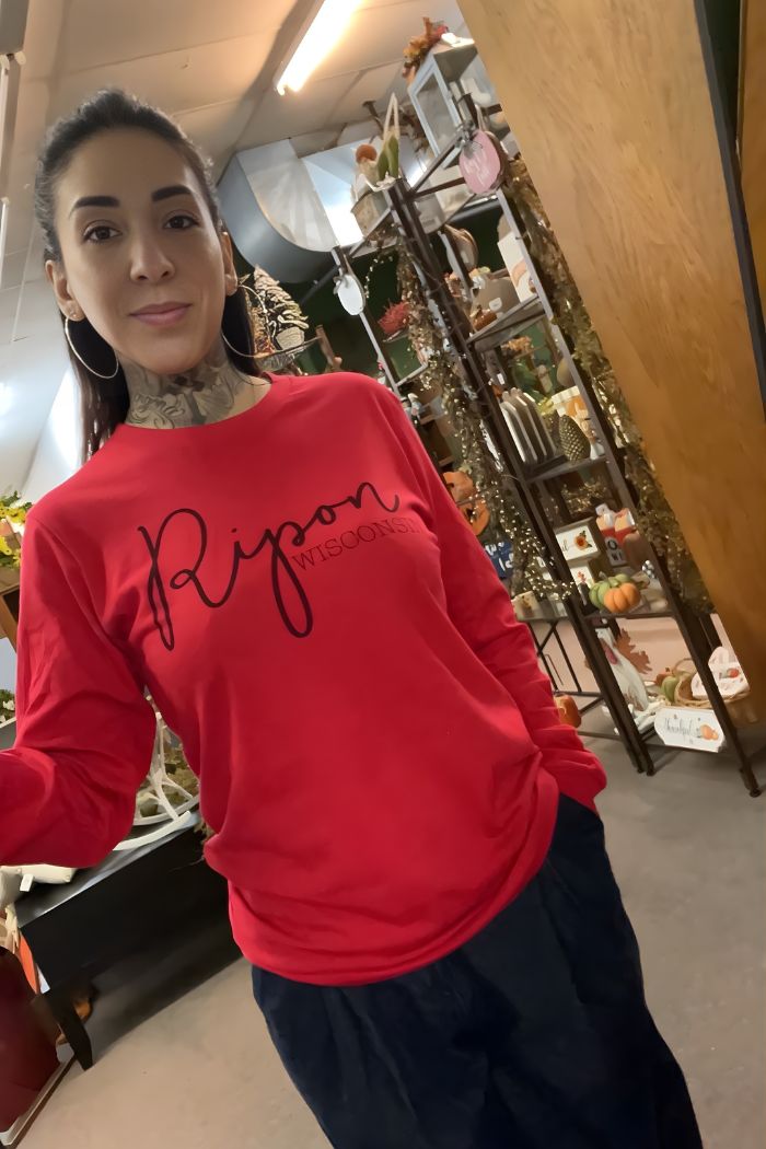 A woman with long dark hair and neck tattoos wears the Ripon Wisconsin Graphic Tee in red with dark pants, standing indoors near shelves decorated with plants and other items.