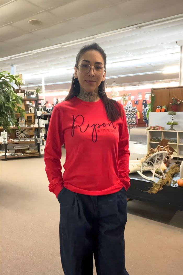 A woman with glasses and tattoos smiles indoors, wearing a bright red Ripon Wisconsin Graphic Tee and dark pants. She stands in a store decorated with shelves, plants, and autumn decor.
