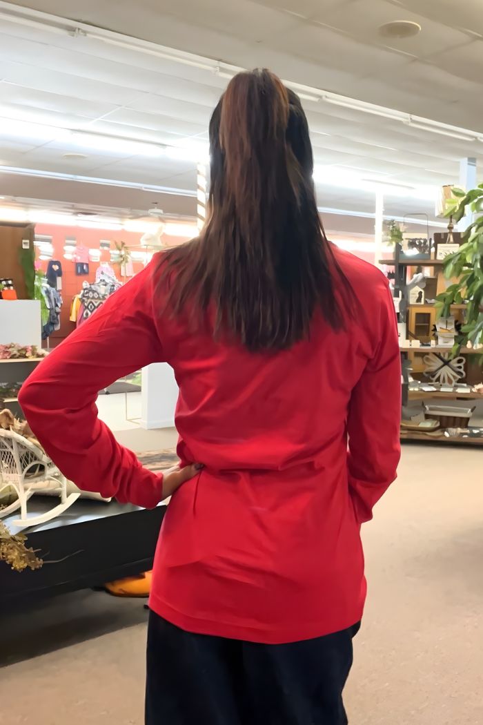 A person with long, dark hair in a ponytail stands indoors, facing away, wearing a bright red Ripon Wisconsin Graphic Tee and dark pants. Store shelves and plants are visible in the background.