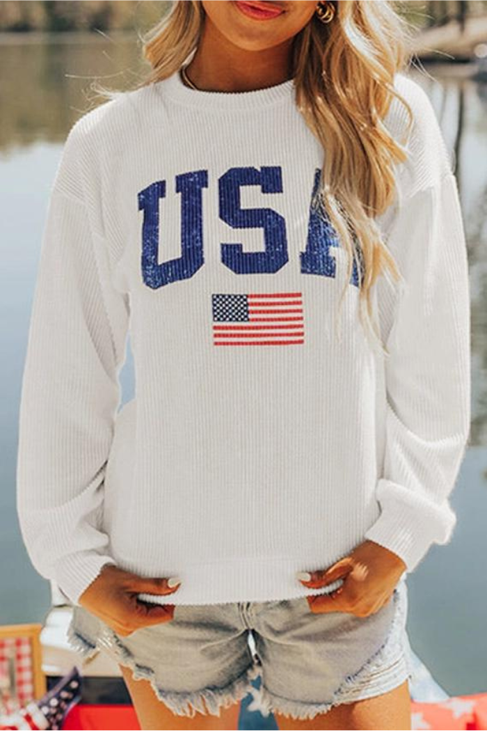 A woman stands outdoors by the water, smiling with her hands in her pockets, wearing a white USA Flag Graphic Sweatshirt featuring blue letters and an American flag, paired with light denim shorts.