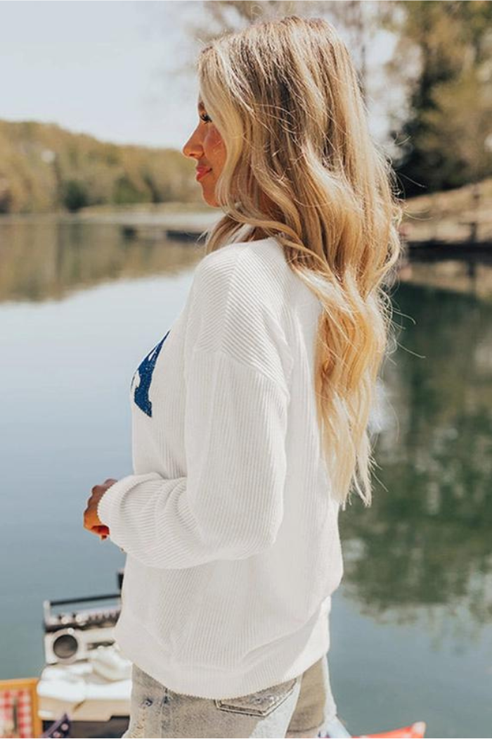 A woman with long, wavy blonde hair stands in profile by a lake on a sunny day, wearing the USA Flag Graphic Sweatshirt and light denim shorts. Trees and water are visible in the background.