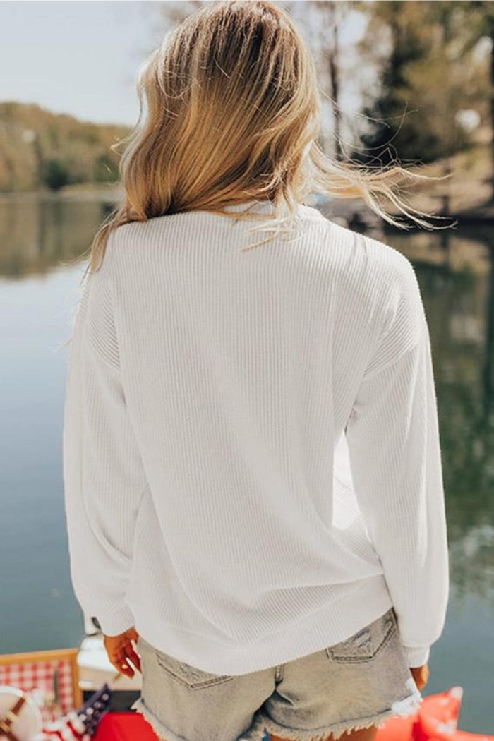 A woman with long, wavy blonde hair stands outdoors by a lake, facing away from the camera. She is wearing a USA Flag Graphic Sweatshirt and light denim shorts, with trees and water visible in the background.