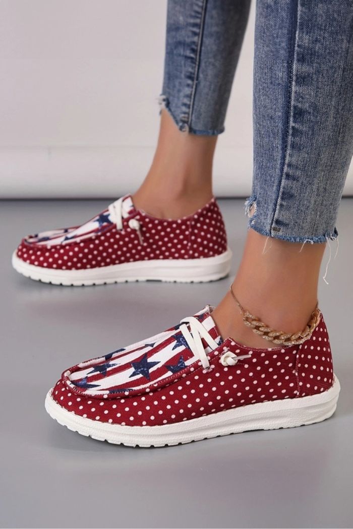 Someone sports the Americana Mash Print Sneakers—red canvas slip-ons with white polka dots, stars-and-stripes accents, and white soles—styled casually with frayed blue jeans and an anklet for a fun, laid-back vibe.