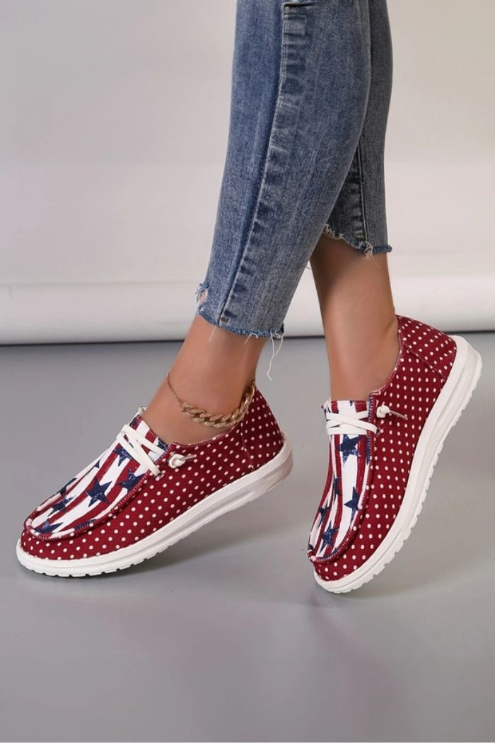 Close-up of a person in blue skinny jeans and an ankle bracelet, showcasing the Americana Mash Print Sneakers with red canvas, white polka dots, and a stars-and-stripes design for a patriotic look.