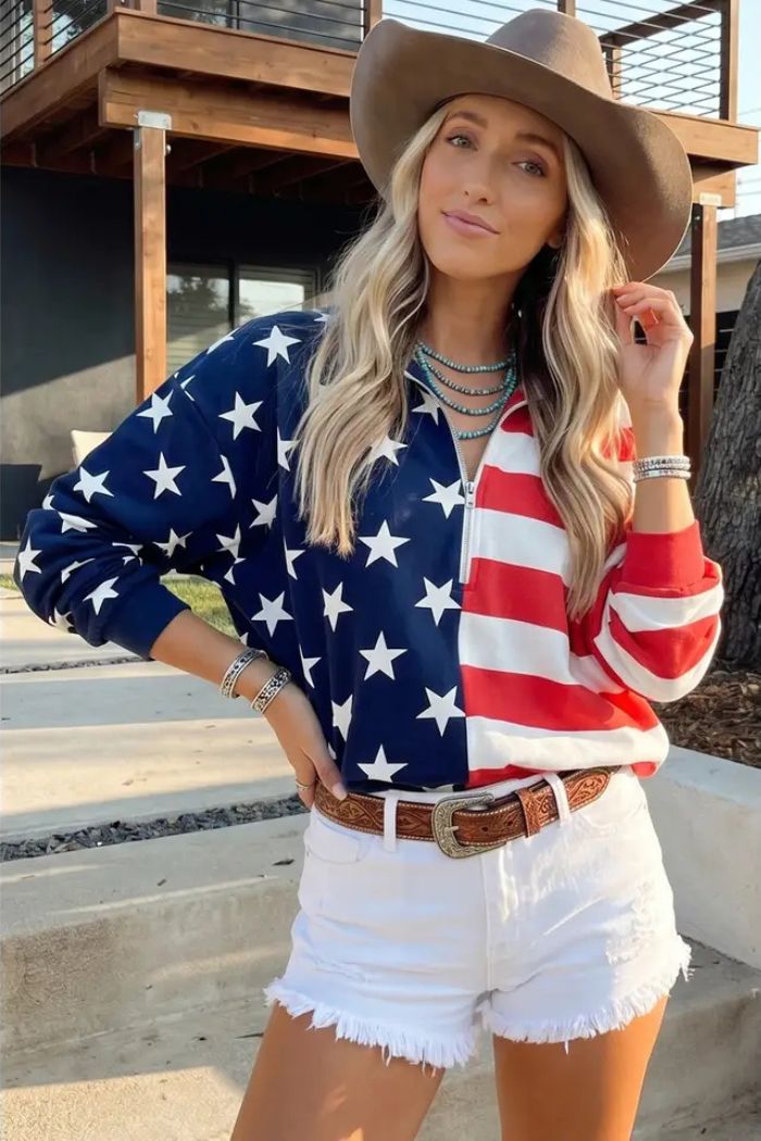 A woman in a cowboy hat wears the USA Flag Quarter Zip with a star-spangled design and white frayed shorts, standing outdoors with one hand on her hip and a relaxed expression.
