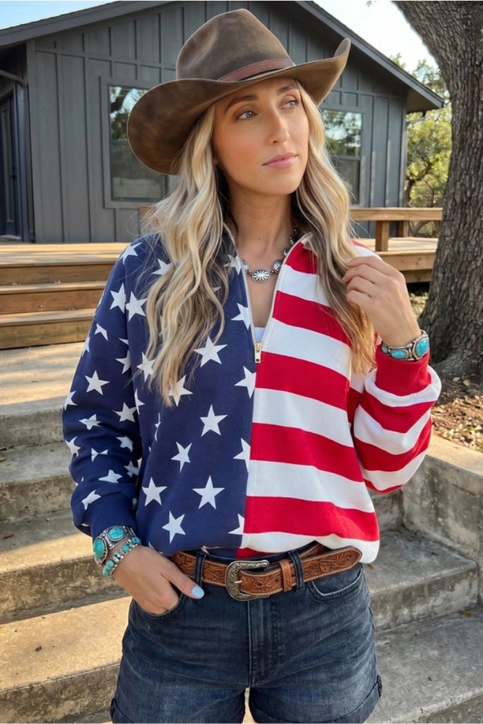 A woman in a cowboy hat wears the USA Flag Quarter Zip and denim shorts outdoors in front of a house, with long wavy blonde hair and turquoise jewelry.