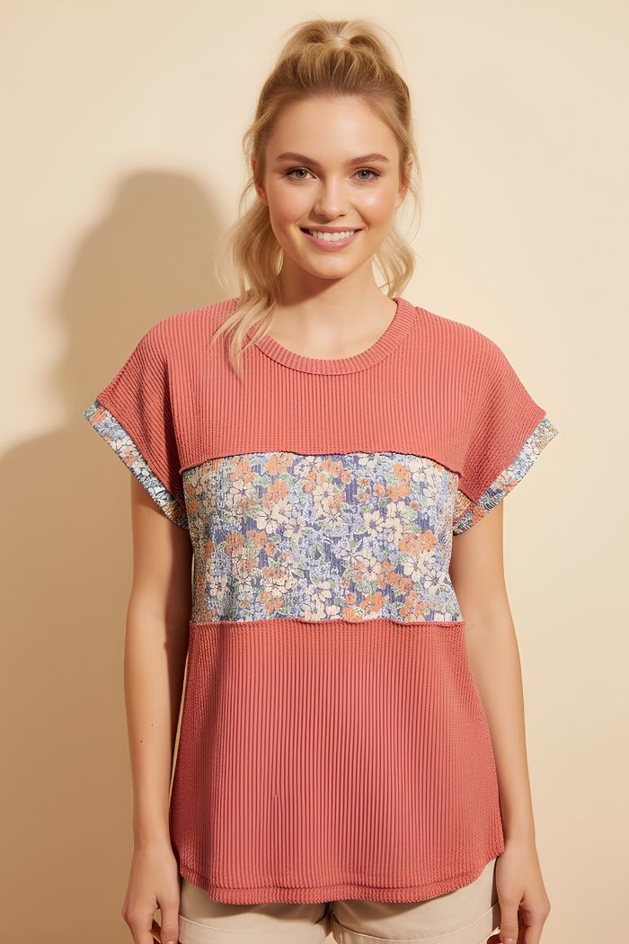A woman with blonde hair in a high ponytail smiles at the camera, wearing a Floral Contrast Top—a short-sleeved, rose knit top with a floral panel—standing against a beige background.