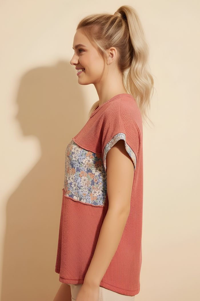 A smiling woman with blonde hair in a ponytail stands in profile against a beige background, wearing the Floral Contrast Top—a relaxed, short-sleeved knit top featuring a floral patterned panel.