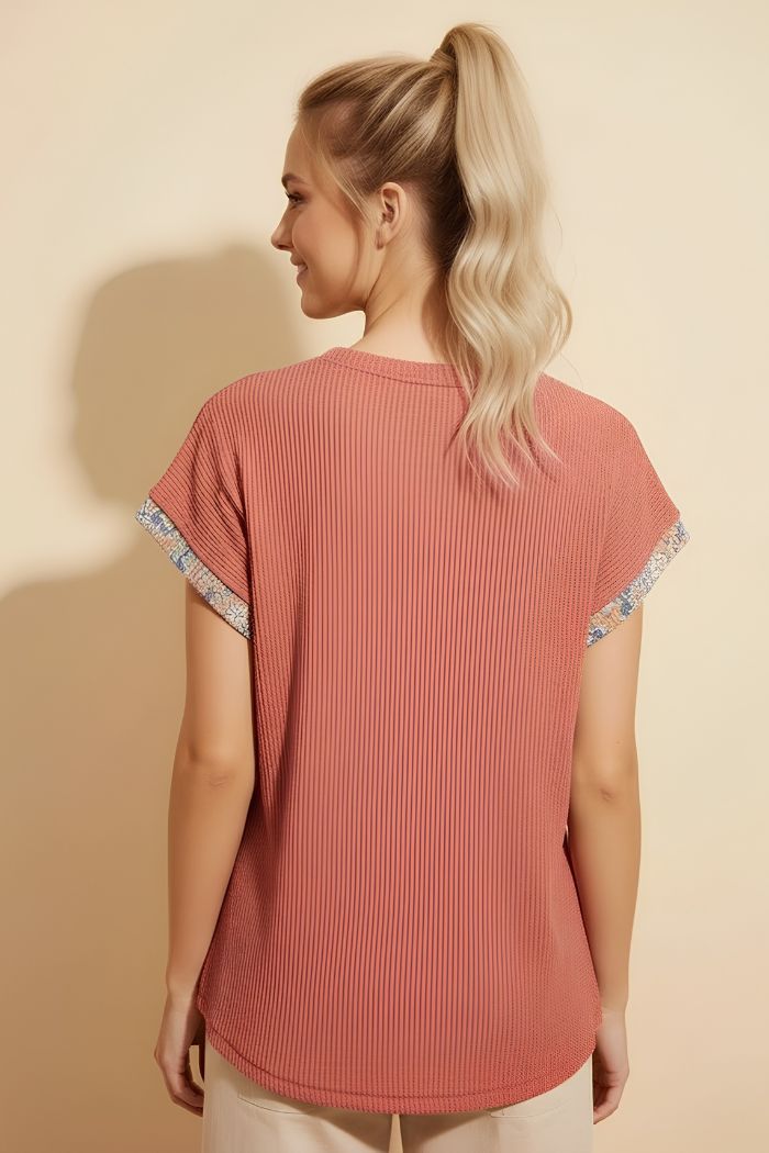 A woman with long blonde hair in a ponytail stands facing away, wearing a relaxed Floral Contrast Top, featuring patterned sleeve trim, against a plain beige background.