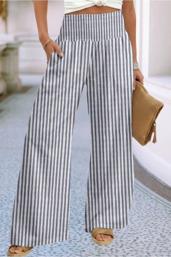 Someone wears Striped Wide Leg Pants in blue and white, a white top, tan sandals, and carries a tan textured clutch bag while standing on a patterned indoor floor.